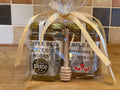Two jars of honey and a honey drizzler in a clear cellophane bag with yellow and purple ribbons on a neutral background.