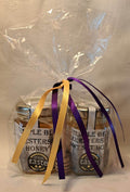 Two jars of honey and a honey drizzler in a clear cellophane bag with yellow and purple ribbons on a neutral background.